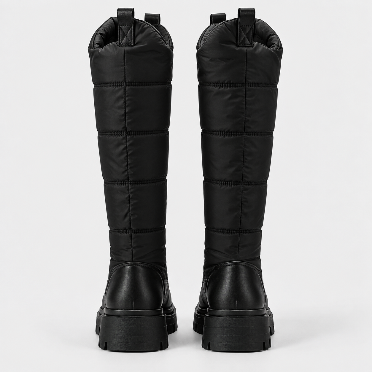 A pair of black quilted knee-high boots with chunky lug soles, shown from the back against a light gray studio background.
