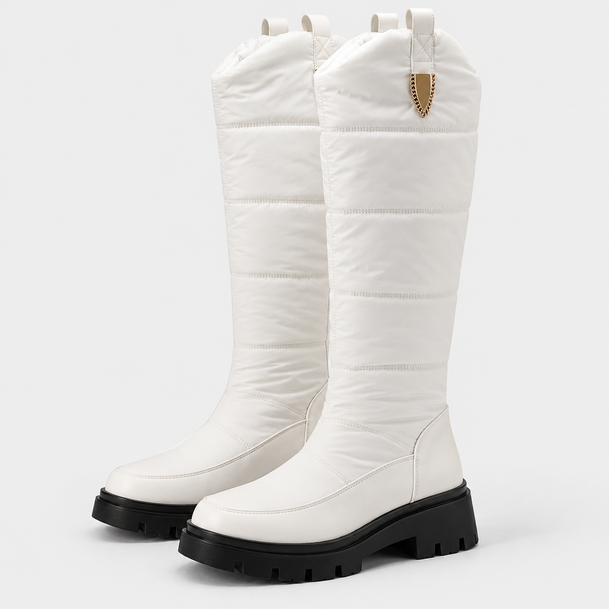 A pair of tall, off-white quilted winter boots with chunky black lug soles and a small gold shield-shaped metal accent on the pull tab, shown against a plain light gray background.