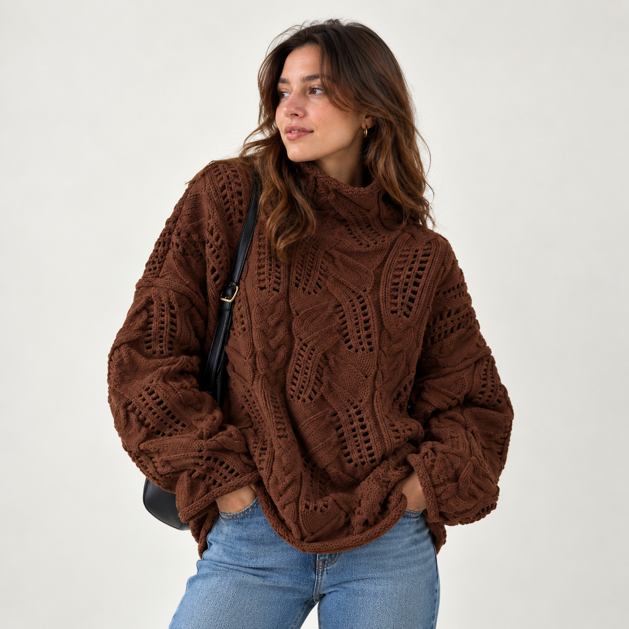 A woman wearing a brown cable-knit mock neck sweater with blue jeans, standing against a plain light gray background.