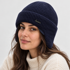 A woman wearing a navy blue ribbed knit beanie with a small dark green logo patch on the front, paired with a cream-colored knit sweater against a light gray background.