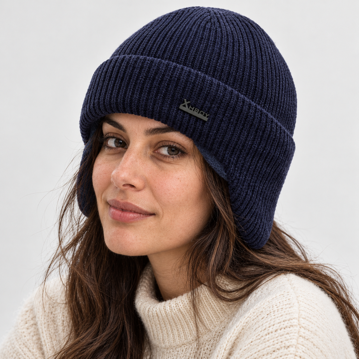 A woman wearing a navy blue ribbed knit beanie with a small dark green logo patch on the front, paired with a cream-colored knit sweater against a light gray background.