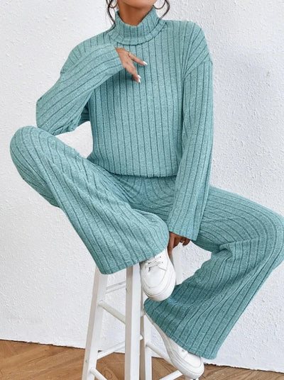 Person wearing a teal ribbed sweater and pants sitting on a white stool against a white wall.