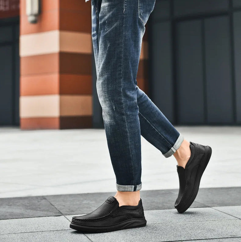 Person wearing blue jeans and black slip-on shoes walking on a sidewalk.