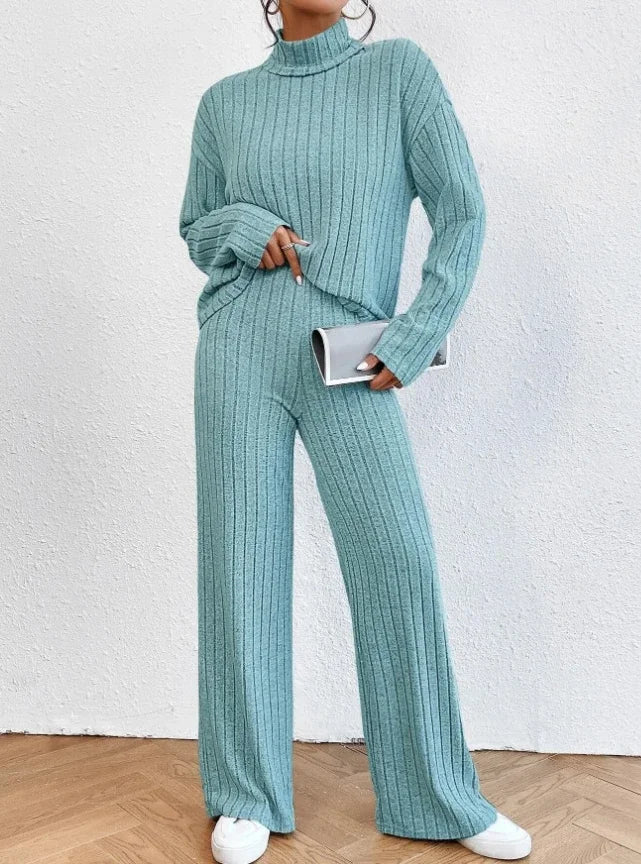 Person wearing a teal ribbed sweater and pants set against a white wall.