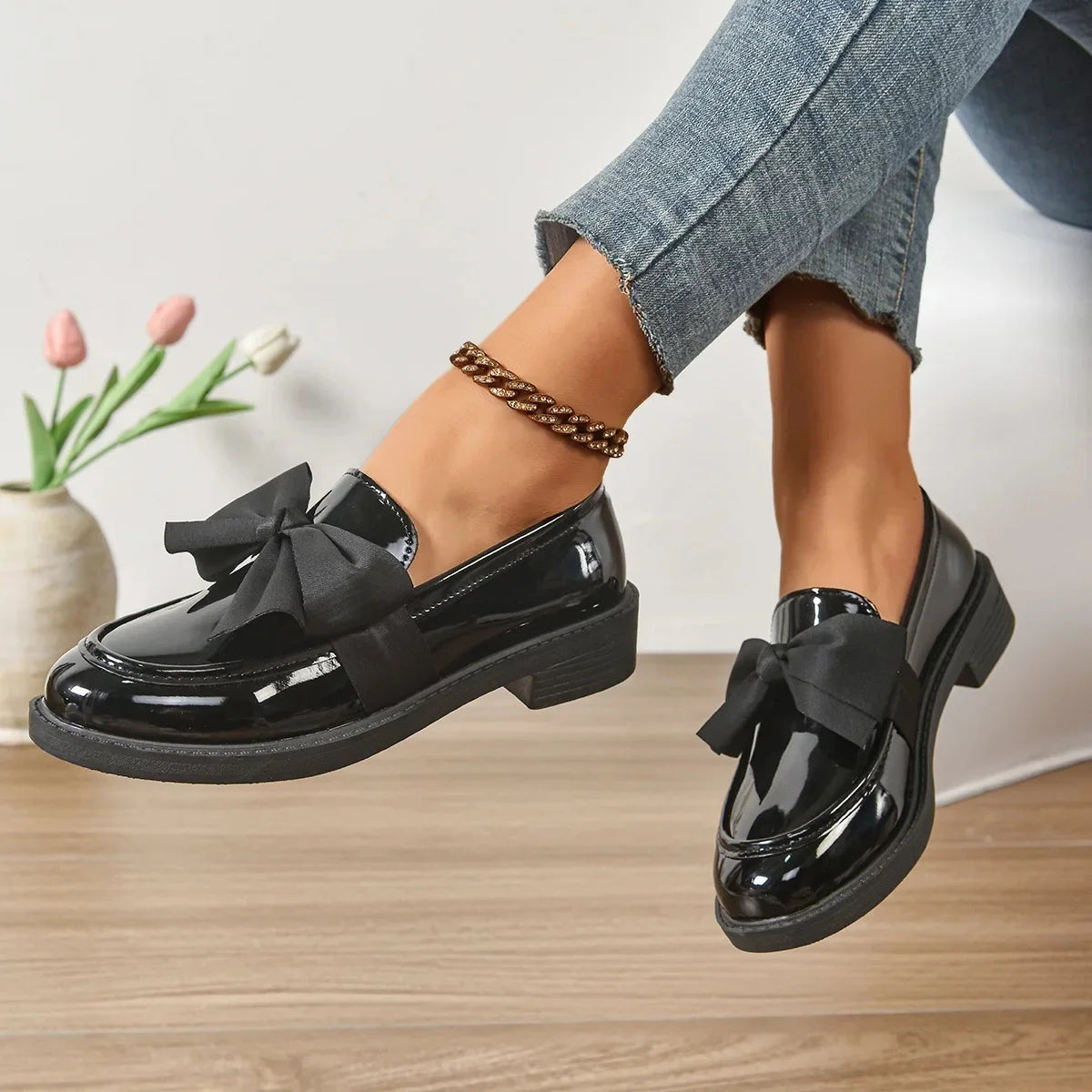 Black patent leather shoes with bows worn by a person sitting on a wooden floor.
