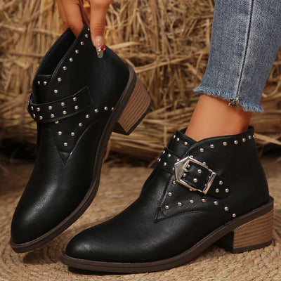 Black studded ankle boots with wooden soles worn by a person against a straw background