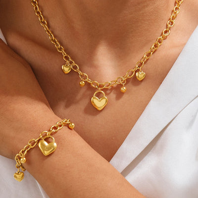 Gold necklace and bracelet with heart and lock charms on a person wearing a white top.