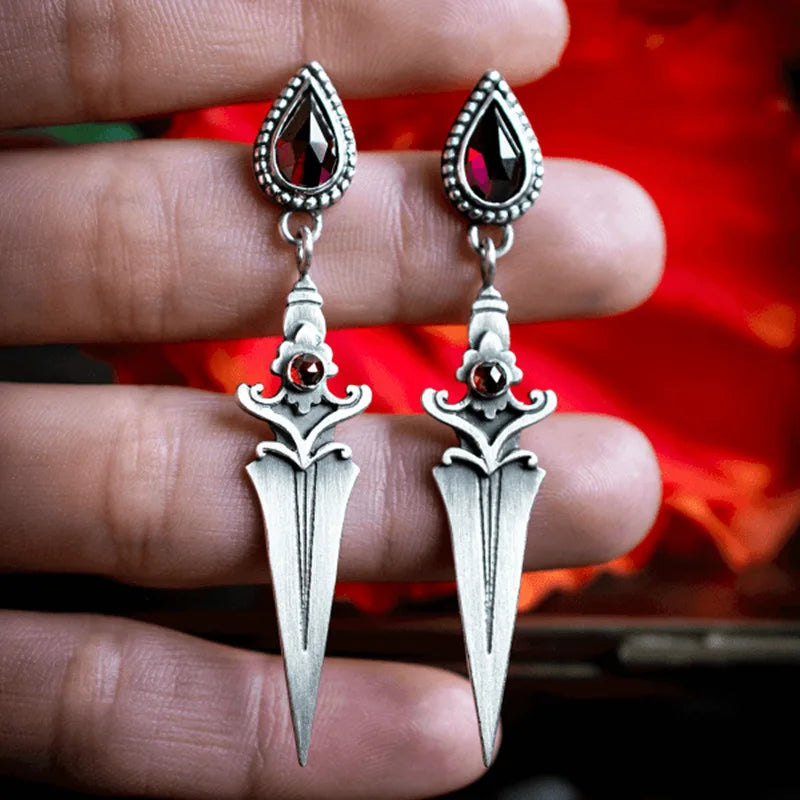 Silver dagger-shaped earrings with red gemstones held between fingers against a blurred background