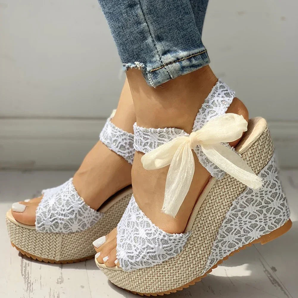 White lace wedge sandals with a bow worn with rolled-up jeans on a light wooden floor.