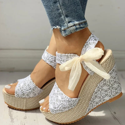 White lace wedge sandals with a bow worn with rolled-up jeans on a light wooden floor.
