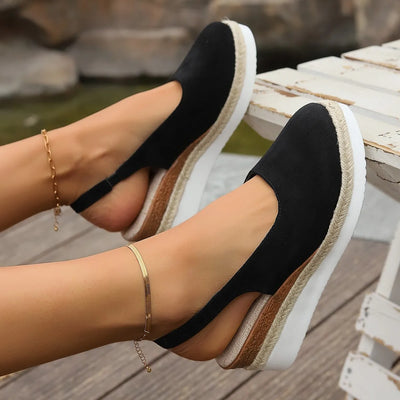 Black wedge sandals with white soles worn by a person on a wooden deck.