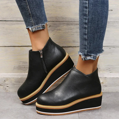 Black wedge ankle boots with brown soles worn with blue jeans on a wooden floor.