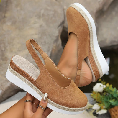 Brown espadrille wedge sandals with white soles held by a person against a blurred natural background.