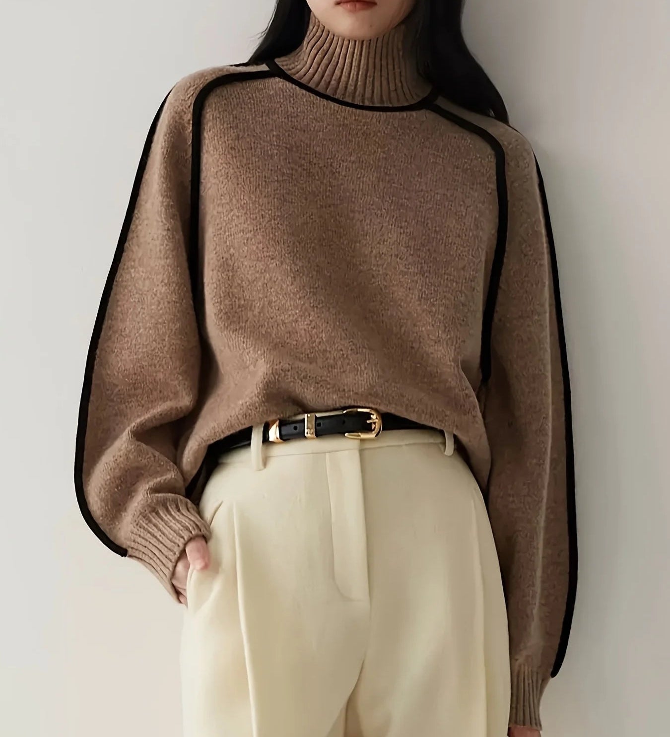Person wearing a brown sweater with black trim and beige pants against a white background