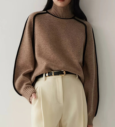Person wearing a brown sweater with black trim and beige pants against a white background