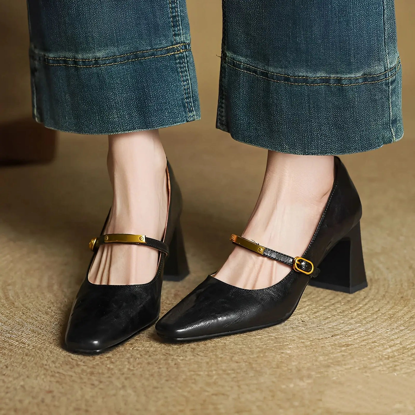 Black high-heeled shoes with gold straps worn with blue jeans on a textured surface.