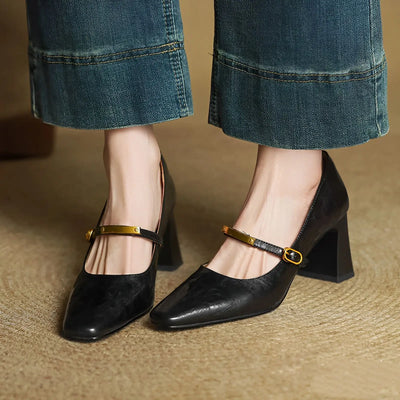 Black high-heeled shoes with gold straps worn with blue jeans on a textured surface.