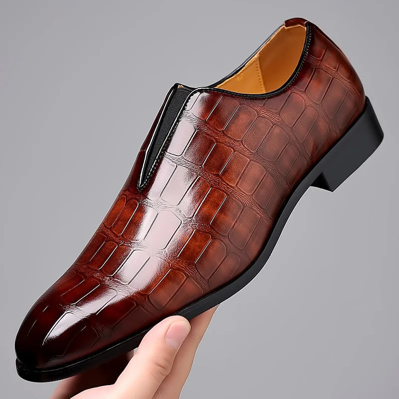Brown leather dress shoe with crocodile pattern held by a hand against a gray background