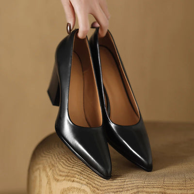 Black high-heeled shoes held by a hand on a wooden surface with a brown background