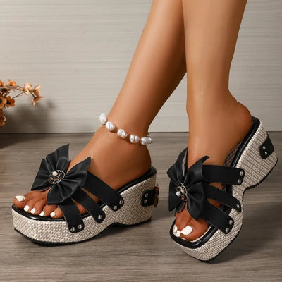 Black platform sandals with bows worn by a person on a wooden floor.