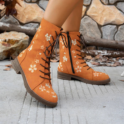 Orange ankle boots with floral embroidery worn by a person on a stone pavement.