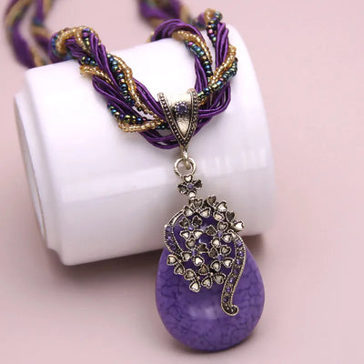 Purple and gold necklace with a decorative pendant on a white stand against a pink background