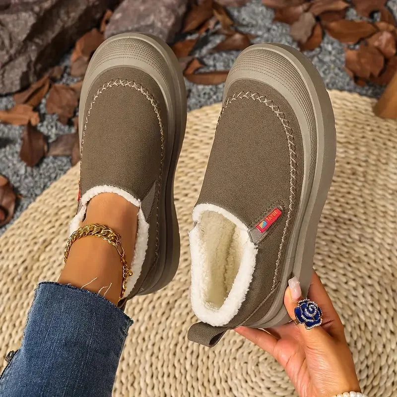 Brown slip-on shoes with white fur lining held by a person against a textured surface.