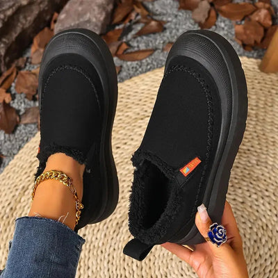 Black slip-on shoes with orange tag held by a hand on a textured surface with leaves.