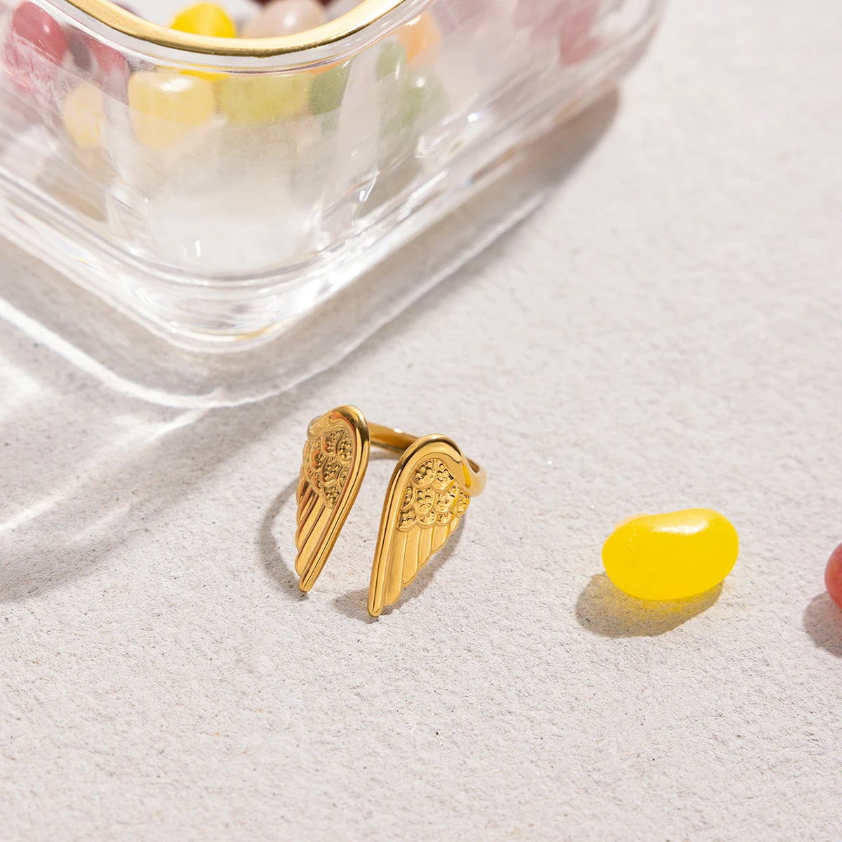 Gold wing-shaped earrings on a white surface with a yellow jelly bean.
