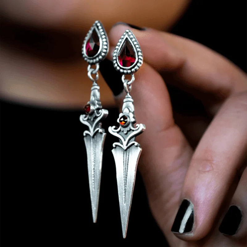 Silver dagger-shaped earrings with red gemstones held by a hand.