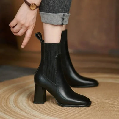 Black leather ankle boots worn with rolled-up jeans on a wooden floor.