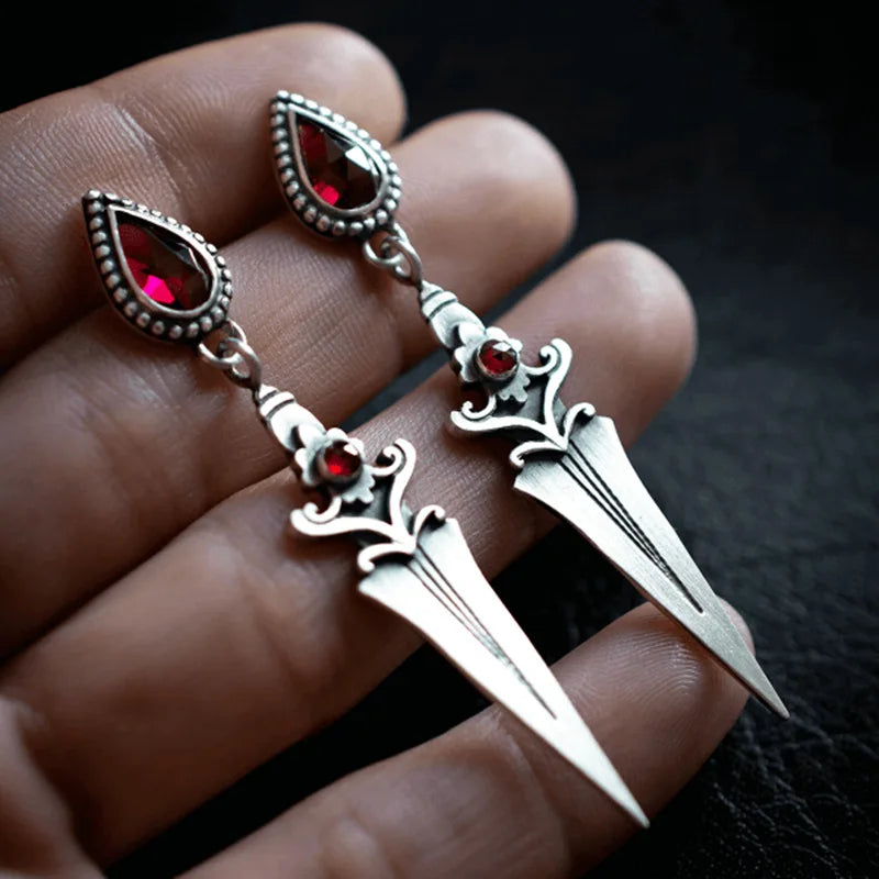 Silver dagger earrings with red gemstones held between fingers against a dark background