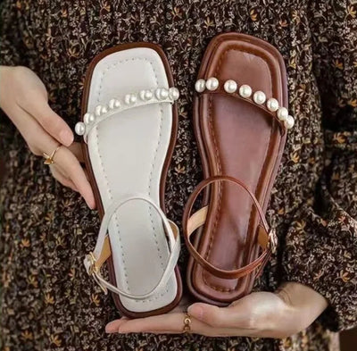 Pair of sandals with pearl embellishments held by a hand on a patterned fabric background