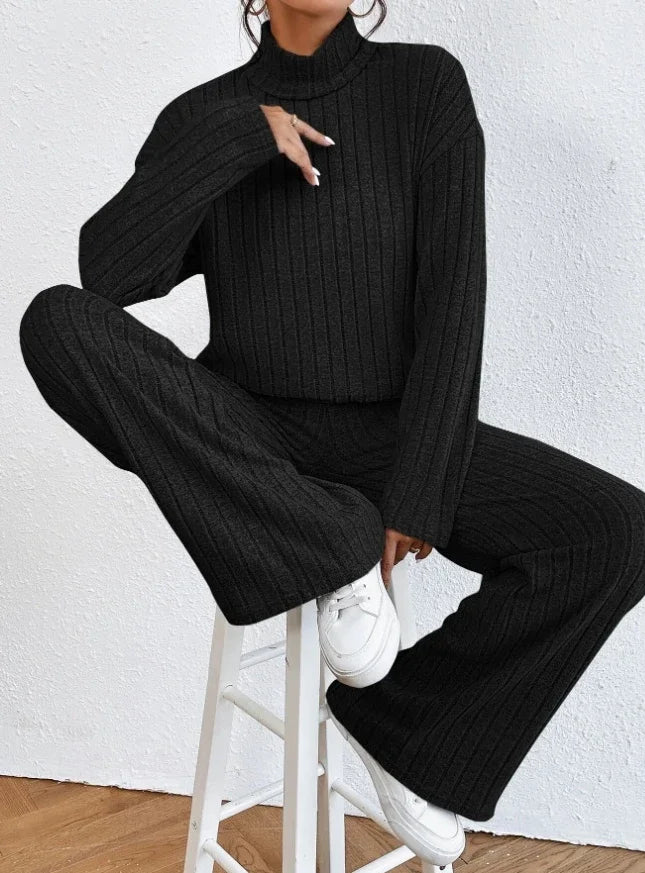 Person wearing a black ribbed sweater and pants sitting on a white stool against a white wall.
