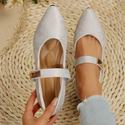 White pointed-toe shoes with a strap held by a hand on a woven mat background