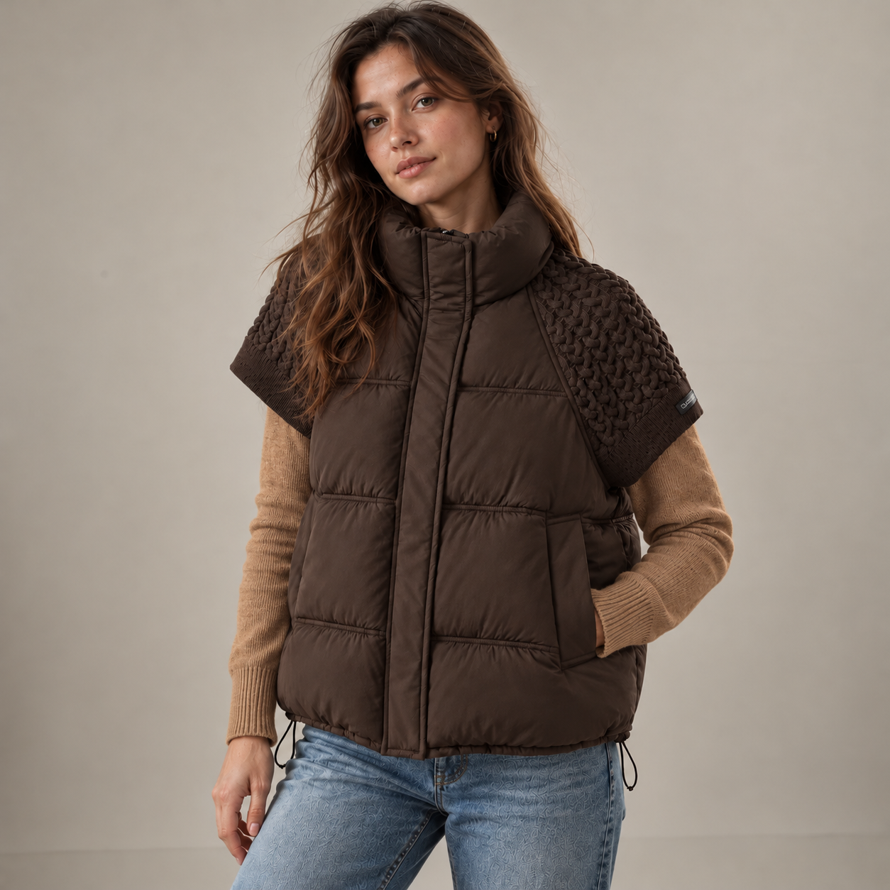 A woman wearing a brown quilted puffer vest with textured knit shoulder panels, layered over a tan long-sleeve sweater and blue jeans, photographed against a plain light gray studio background.