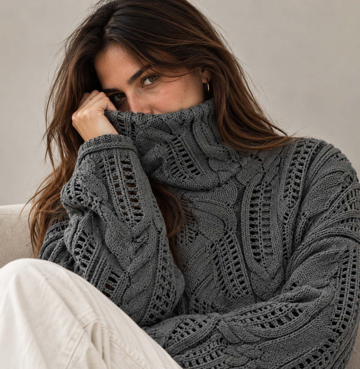 A woman wearing a chunky, dark gray cable-knit turtleneck sweater, holding the collar up near her face. She is also wearing light beige pants, seated against a neutral, light gray background.