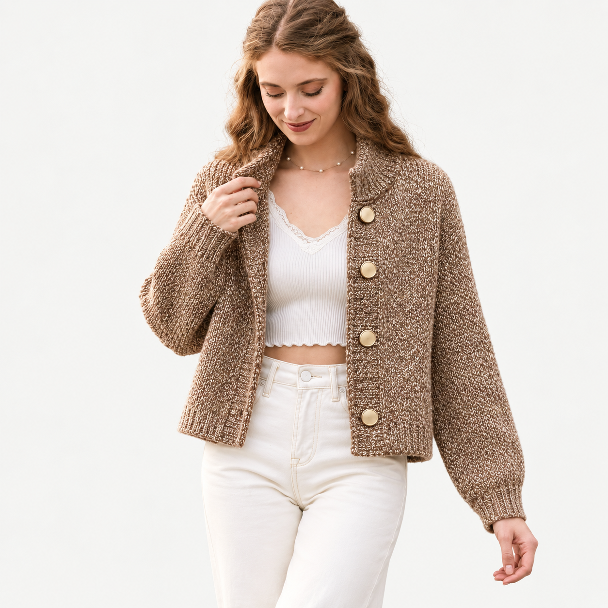 A woman wearing a brown marled knit cardigan with large gold buttons, styled over a white ribbed crop top and white jeans, against a plain white background.