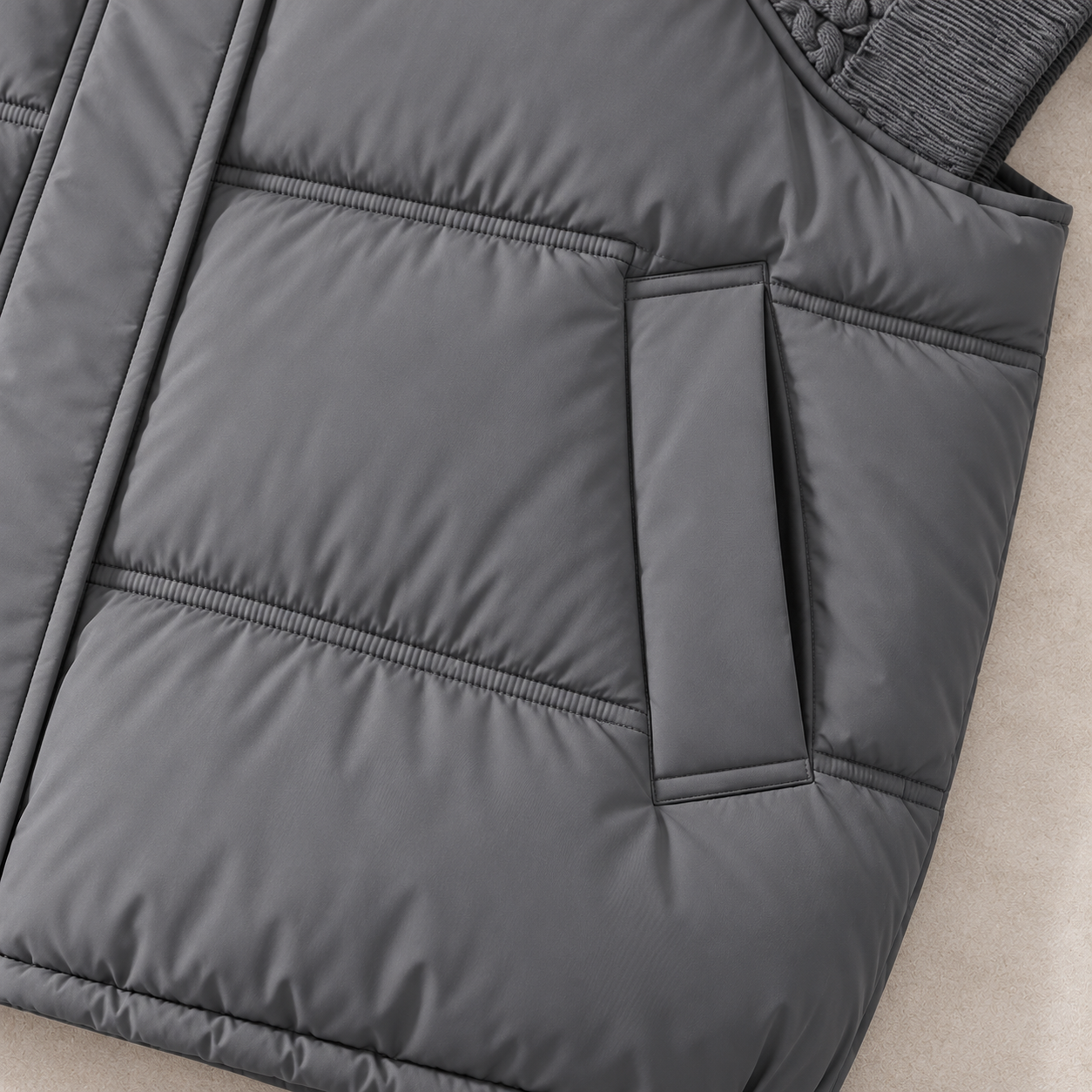 Close-up of a dark gray quilted puffer vest showing the padded paneling, zipper pocket, and ribbed knit collar on a neutral beige surface.